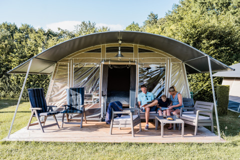 A family is relaxing outside a modern tent lodge, sitting on outdoor chairs and enjoying the sunny weather.