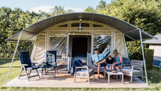 Eine Familie entspannt sich vor einer modernen Zelt-Lodge mit Sitzgelegenheiten und Tisch im Grünen.