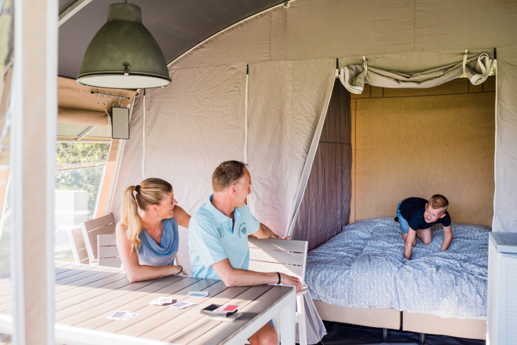 Een gezin geniet in een moderne lodge; ouders zitten aan tafel terwijl een kind op het bed aan het spelen is.