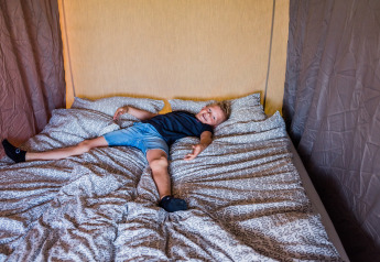 A child is sprawled out on a patterned bed in a cozy lodge room, smiling happily at the camera.