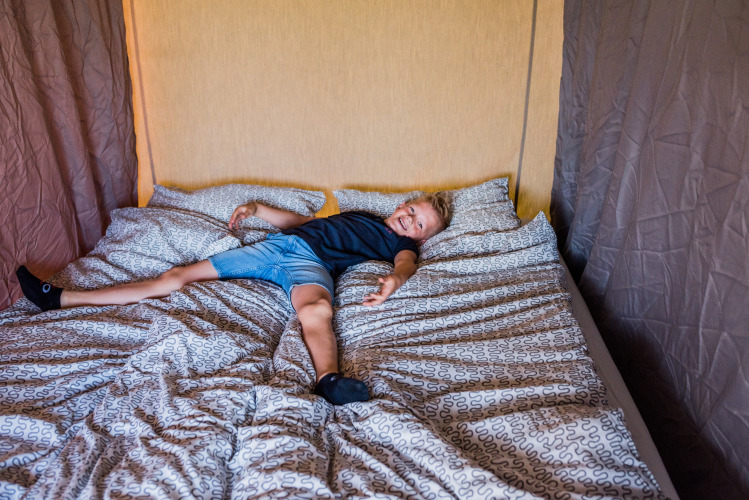 A child is sprawled out on a patterned bed in a cozy lodge room, smiling happily at the camera.