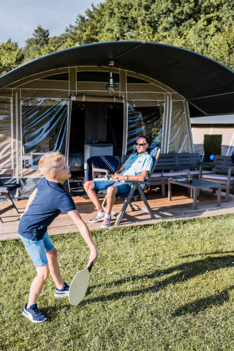 Boy playing badminton on the grass outside a luxury lodge while a man relaxes on the patio behind him.