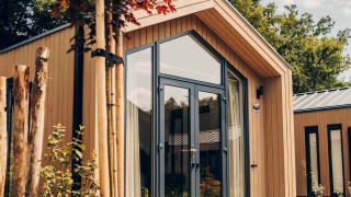 Tiny Lodge moderne aux grandes baies vitrées, jardin naturel, sous un ciel ensoleillé et entouré d’arbres.