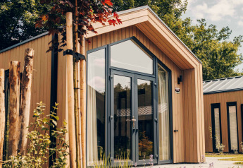 Modernes Tiny House Tiny Lodge mit großen Fenstern, umgeben von Pflanzen und Bäumen an einem sonnigen Tag.