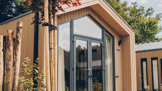 Moderne Tiny Lodge med store vinduer og naturlig have, solbeskinnet dag med træer i baggrunden.