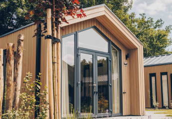 Tiny Lodge moderne aux grandes baies vitrées, jardin naturel, sous un ciel ensoleillé et entouré d’arbres.