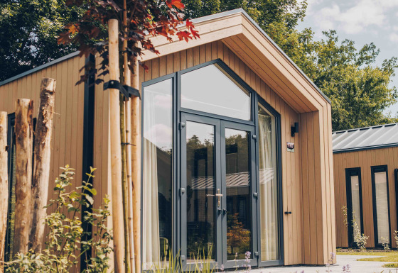 Tiny Lodge met grote ramen, lichte gevelbekleding, tuin met planten en bomen op een zonnige dag.