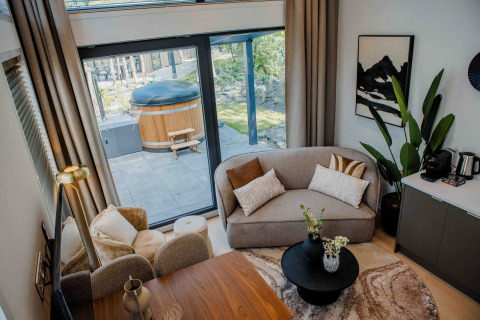 Modern lodge living area with large windows, cozy sofas, stylish decor, and outdoor hot tub visible outside.