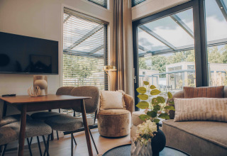 Modernes Lodge-Wohnzimmer mit großen Fenstern, Essbereich, grauem Sofa, TV und Blick ins Grüne.