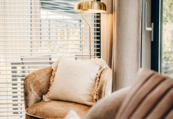 Lyst loungemiljø i en lodge med bløde møbler, en pude, gulvlampe og store vinduer med persienner.