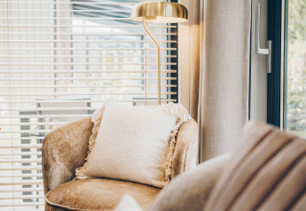 Lyst loungemiljø i en lodge med bløde møbler, en pude, gulvlampe og store vinduer med persienner.