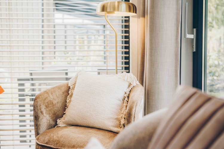 Coin salon lumineux dans un lodge avec fauteuil, coussin, lampe dorée et grandes fenêtres à stores.