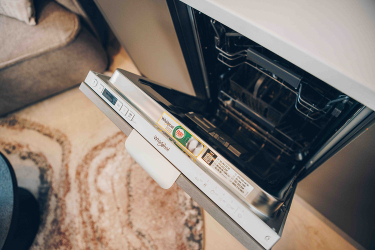 Open Whirlpool dishwasher in a cozy lodge setting with a carpet and sofa visible in the background.