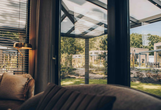 Zicht vanuit een lodge door grote ramen op een moderne buitenruimte met tuin en terrasmeubilair.