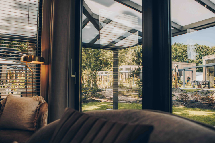 Uitzicht vanuit een lodge-interieur door grote ramen op een modern terras en groene tuin buiten.