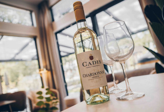 Fles Cadis Chardonnay en twee wijnglazen op een tafel in een gezellige lodge met grote ramen en uitzicht.