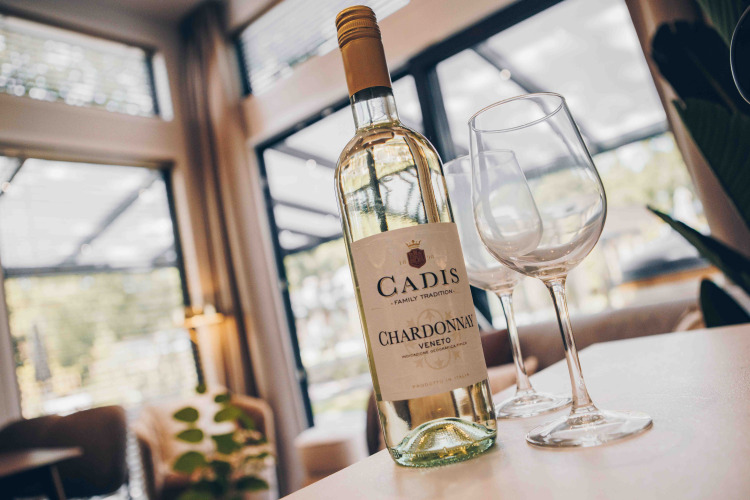 A bottle of Cadis Chardonnay and two empty wine glasses on a table in a cozy lodge with large windows.