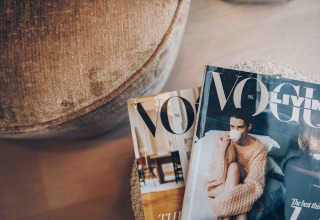 Twee tijdschriften, waaronder Vogue, liggen op een zacht tafeltje bij een stoel in een gezellige lodge.