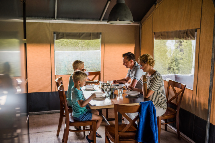 A family of four enjoys a meal together at a table inside a cozy safari tent with natural lighting.