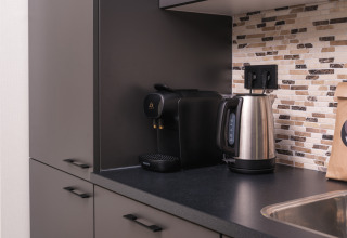 Moderne tekøkken med espressomaskine, elkedel og sort bordplade på et lodge-værelse med flisevæg.