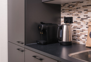 Moderne tekøkken med espressomaskine, elkedel og sort bordplade på et lodge-værelse med flisevæg.