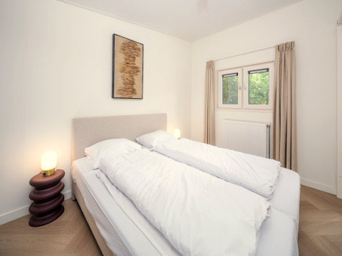 Helles, modernes Schlafzimmer in einer Lodge mit Doppelbett, Lampen, Kunstbild und Fenster mit Vorhängen.