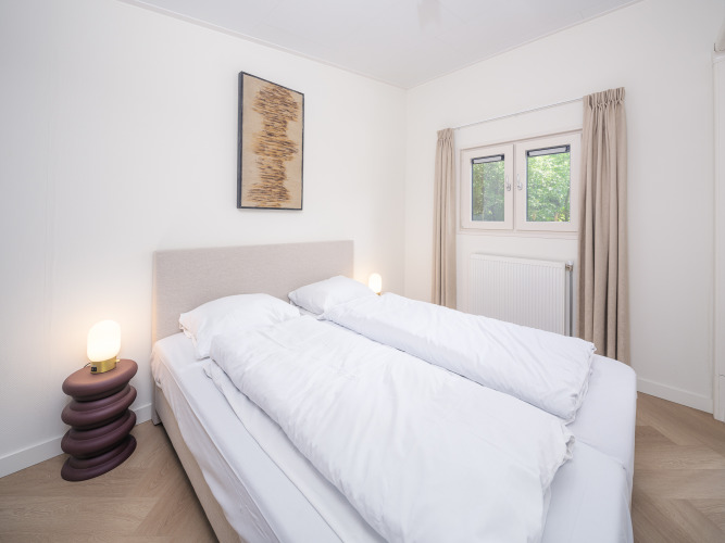 Helles, modernes Schlafzimmer in einer Lodge mit Doppelbett, Lampen, Kunstbild und Fenster mit Vorhängen.