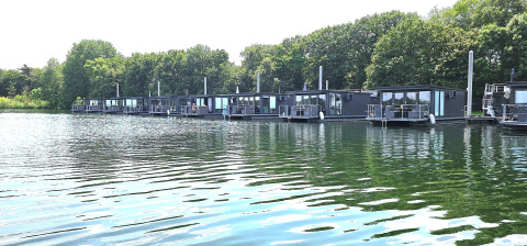 Plusieurs péniches modernes flottent sur un lac paisible, entourées d’arbres verts et d’un ciel clair.