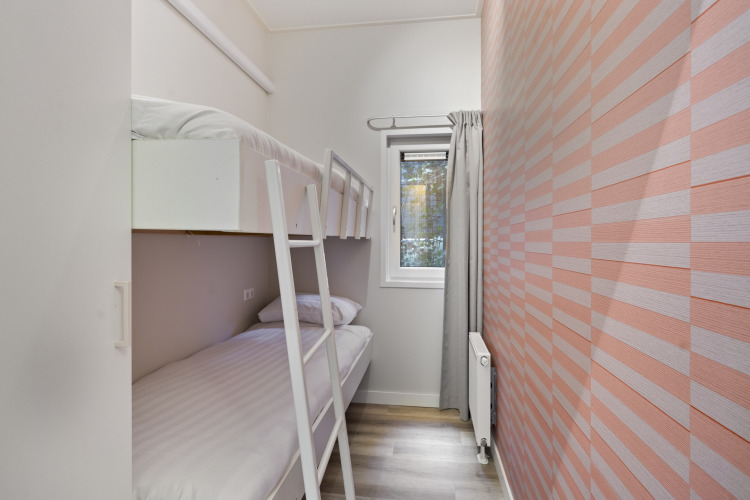 Schmales Zimmer mit Etagenbett, kleinem Fenster, hellem Vorhang und rosa gestreifter Wand im Ferienhaus.