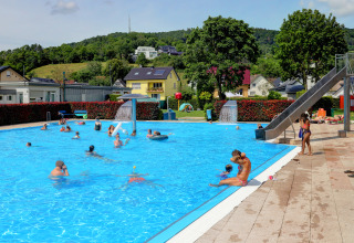 Varias personas disfrutan en la piscina al aire libre de Campingpark Echternacherbrück entre verdes colinas alemanas.