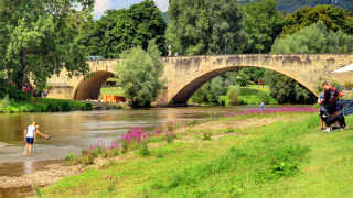 Ponte di pietra su fiume con persone, fiori viola e cane al Campingpark Echternacherbrück, Germania.