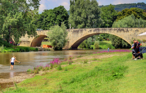Stone bridge over river with people, purple flowers, and dogs at Campingpark Echternacherbrück, Germany.