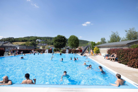 Outdoor swimming pool with guests, sunny weather and green hills at Campingpark Echternacherbrück, Germany.