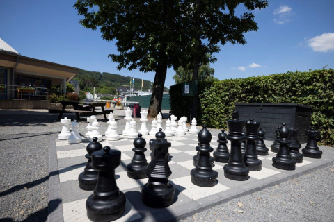 Outdoor giant chess set at Campingpark Echternacherbrück, with trees, benches, and a clear blue sky above.
