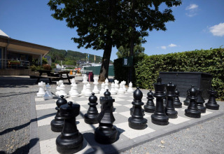 Outdoor giant chess set at Campingpark Echternacherbrück, with trees, benches, and a clear blue sky above.