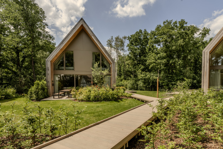 Lodge moderno en Woodz Lodges en Bélgica, rodeado de árboles, jardines y camino de madera bajo el sol.