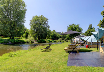 Riverfront view with camping tent, picnic table, and lounge chairs at Campingpark Echternacherbrück, Germany.