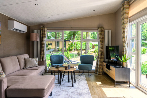 Bright, modern lodge living room with sofa, armchairs, coffee tables, TV, and large windows to greenery.