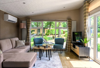 Gezellige, lichte lodge woonkamer met zetel, stoelen, salontafels, tv en groot raam met tuinzicht.