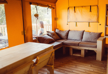 Hyggelig safari-teltinteriør med træmøbler og hjørnesofa, en person sidder udenfor ved Camping Liefrange.