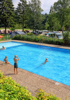 Piscina all'aperto con bambini, lettini prendisole e camper sullo sfondo ad Auf Kengert, Diekirch, Lussemburgo.