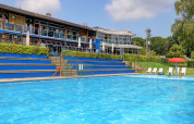 Buitenzwembad aan een vakantiepark in Diekirch, Luxemburg, met ligstoelen en moderne gebouwen op de achtergrond.
