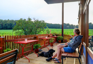 Kvinde og sort hund sidder på en terrasse med udsigt over grønne marker ved Auf Kengert feriepark i Luxembourg.