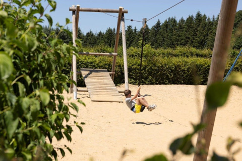 Dreng leger på svævebane på en legeplads omgivet af træer i Auf Kengert-ferieparken, Diekirch, Luxembourg.