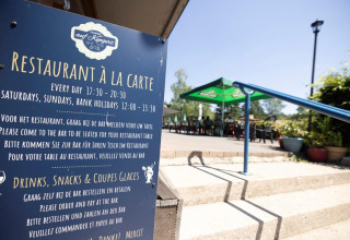 Restaurant-terrasse ved Auf Kengert feriepark i Diekirch, Luxembourg, med informationsskilt og parasoller.