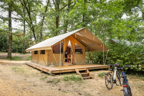 Tienda de safari clásica sobre plataforma de madera en el bosque, dos bicicletas aparcadas delante.