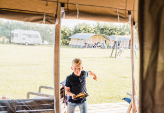 Boy steps into Auf Kengert Lodge de Luxe in Luxembourg, carrying food. Camping tents and caravan outside.