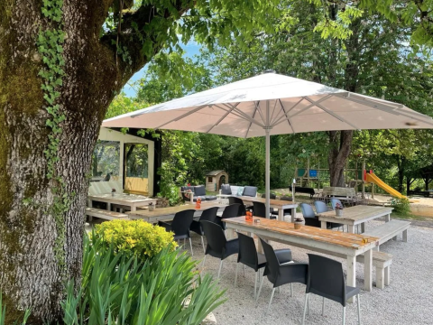 Outdoor dining area at Camping Le Clou featuring large umbrellas, wooden benches, and lush green trees.