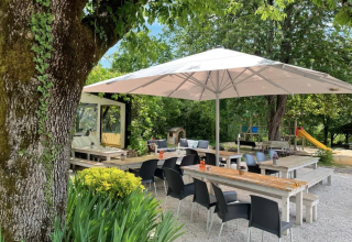 Outdoor dining area at Camping Le Clou featuring large umbrellas, wooden benches, and lush green trees.