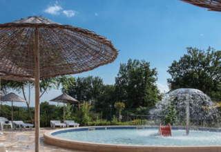 Circular pool with a water fountain and straw parasols at Camping Le Clou, Nouvelle-Aquitaine, France.